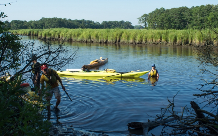 a person stands in waist-deep water beside a couple of floating kayaks, while others navigate a different kayak toward the shore. 